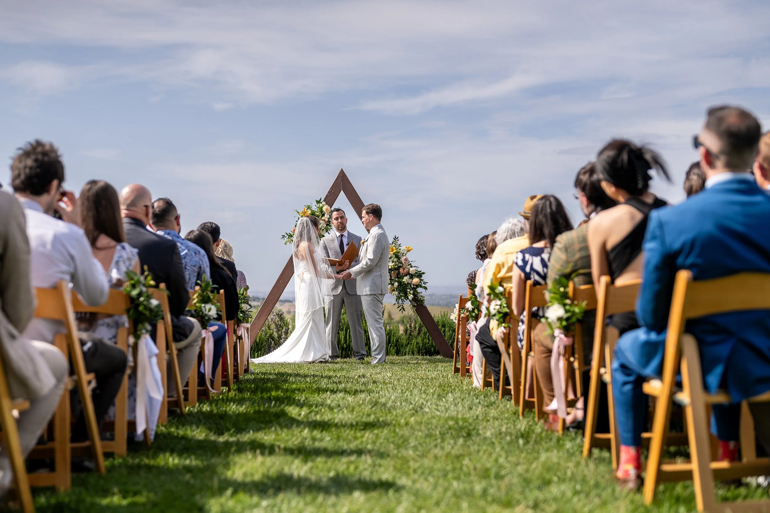 A wedding ceremony taking place outdoors on a grassy field with a blue sky. The bride and groom stand under a wooden triangle arch adorned with flowers, exchanging vows. Guests sit on wooden chairs decorated with greenery, facing the couple.
