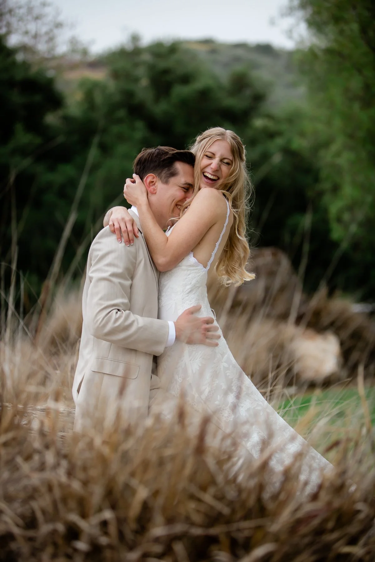 A joyful couple embracing outdoors, with the woman in a white wedding dress and the man in a beige suit, surrounded by tall grass and greenery.