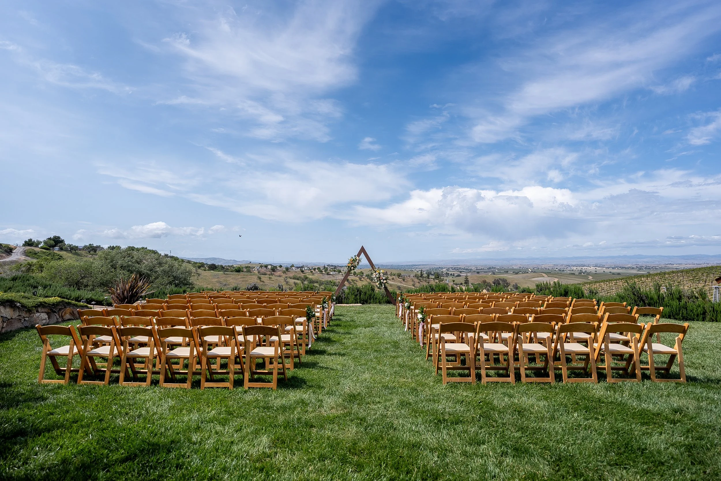 Outdoor wedding ceremony setup with rows of wooden chairs facing a triangular arch decorated with flowers, set on a grassy field with scenic hills under a partly cloudy blue sky.