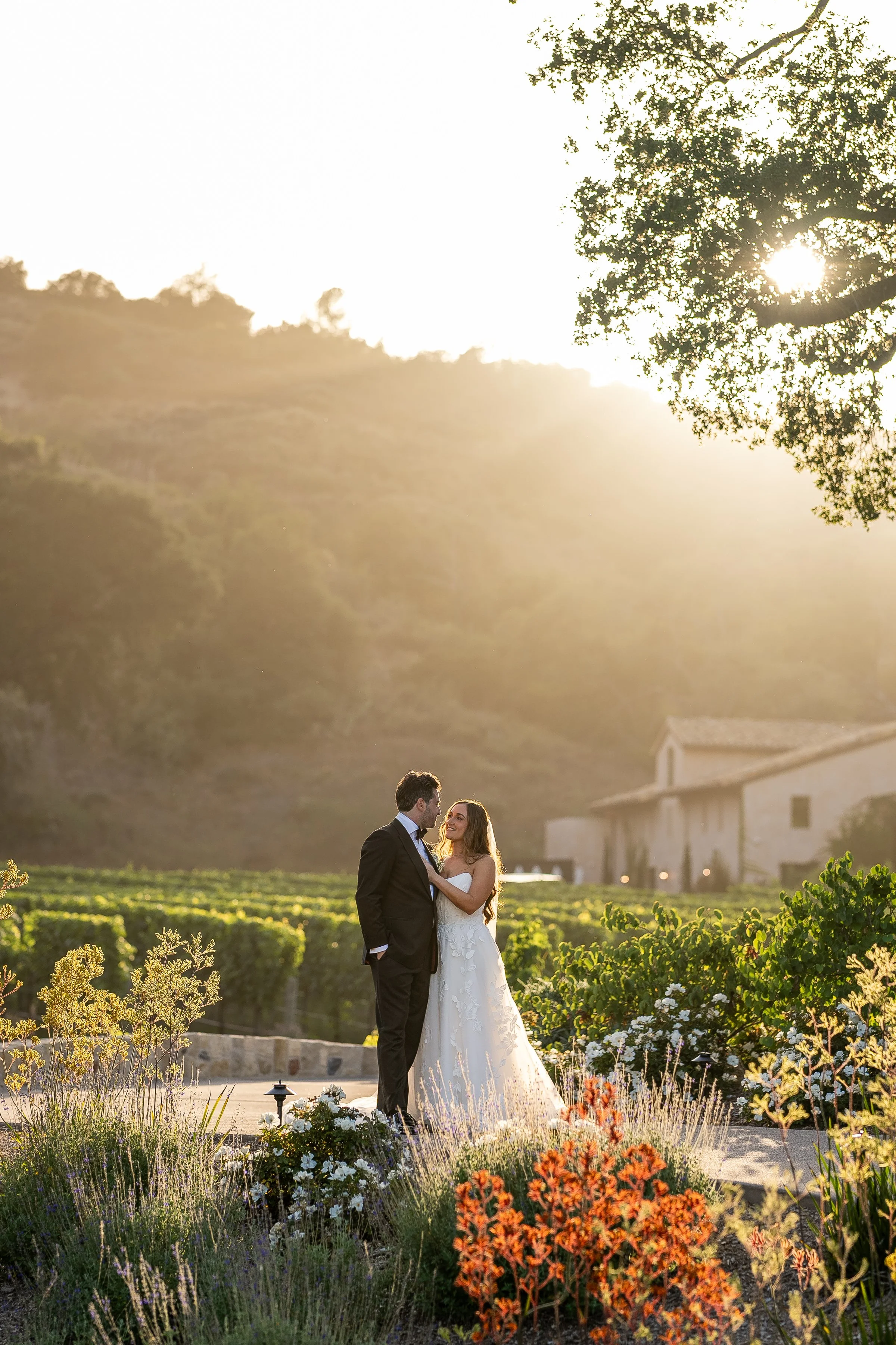 A bride and groom standing together outdoors at sunset, surrounded by flowers and greenery, with a vineyard and hills in the background.