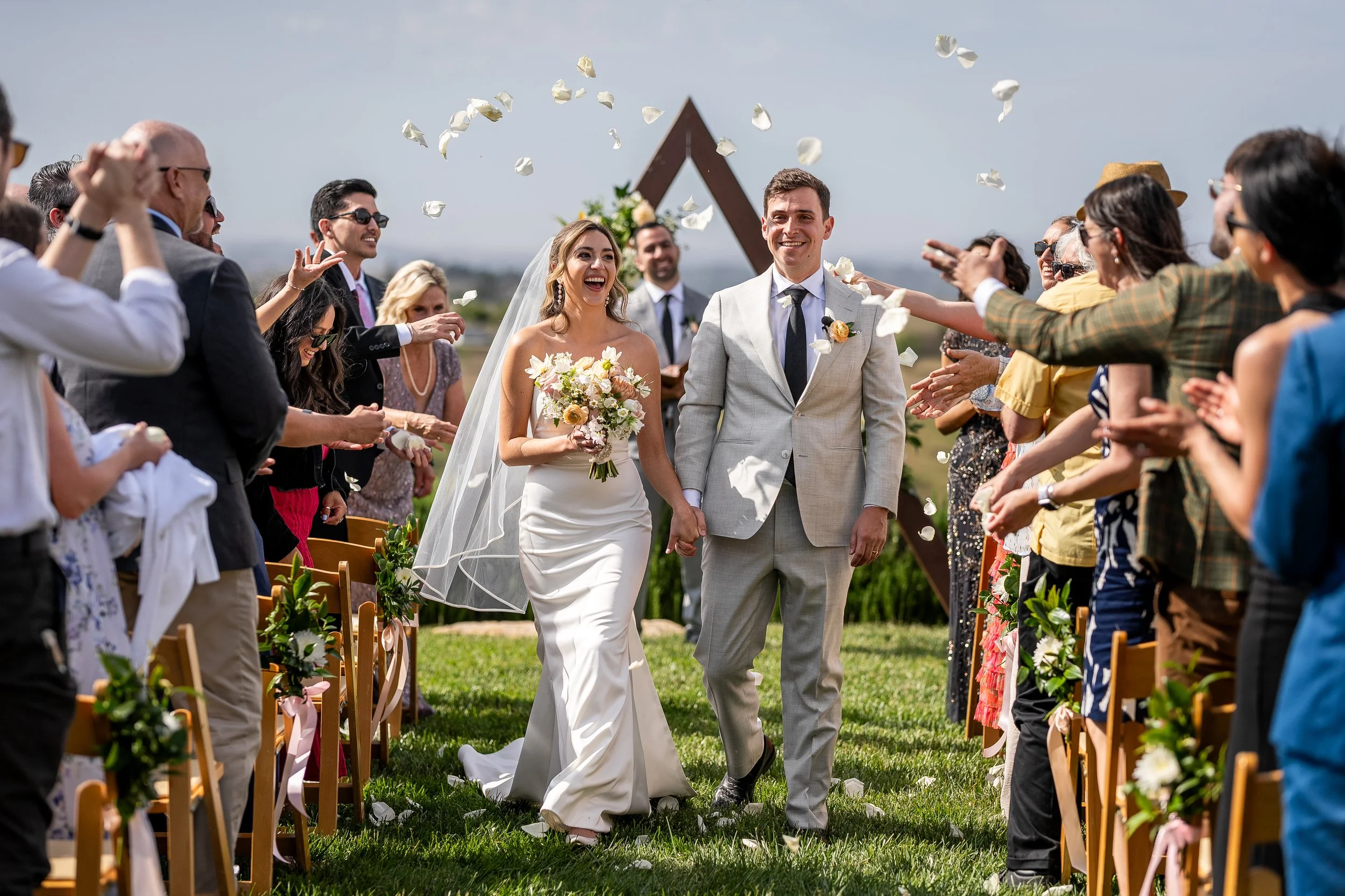 A newlywed couple walks down the aisle after their wedding ceremony, surrounded by friends and family throwing flower petals. The bride is in a white wedding dress holding a bouquet, and the groom is in a light gray suit. The outdoor setting has a cl