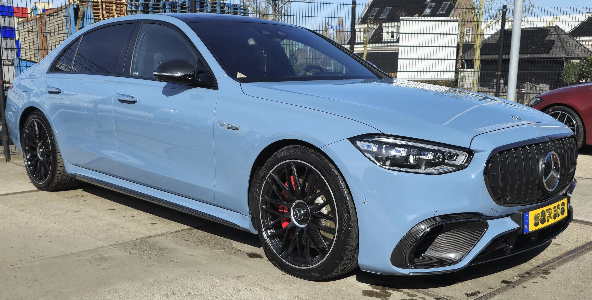 Lichtblauwe luxeauto, mogelijk Mercedes, op een parkeerplaats met zwarte velgen en gele kentekenplaat.