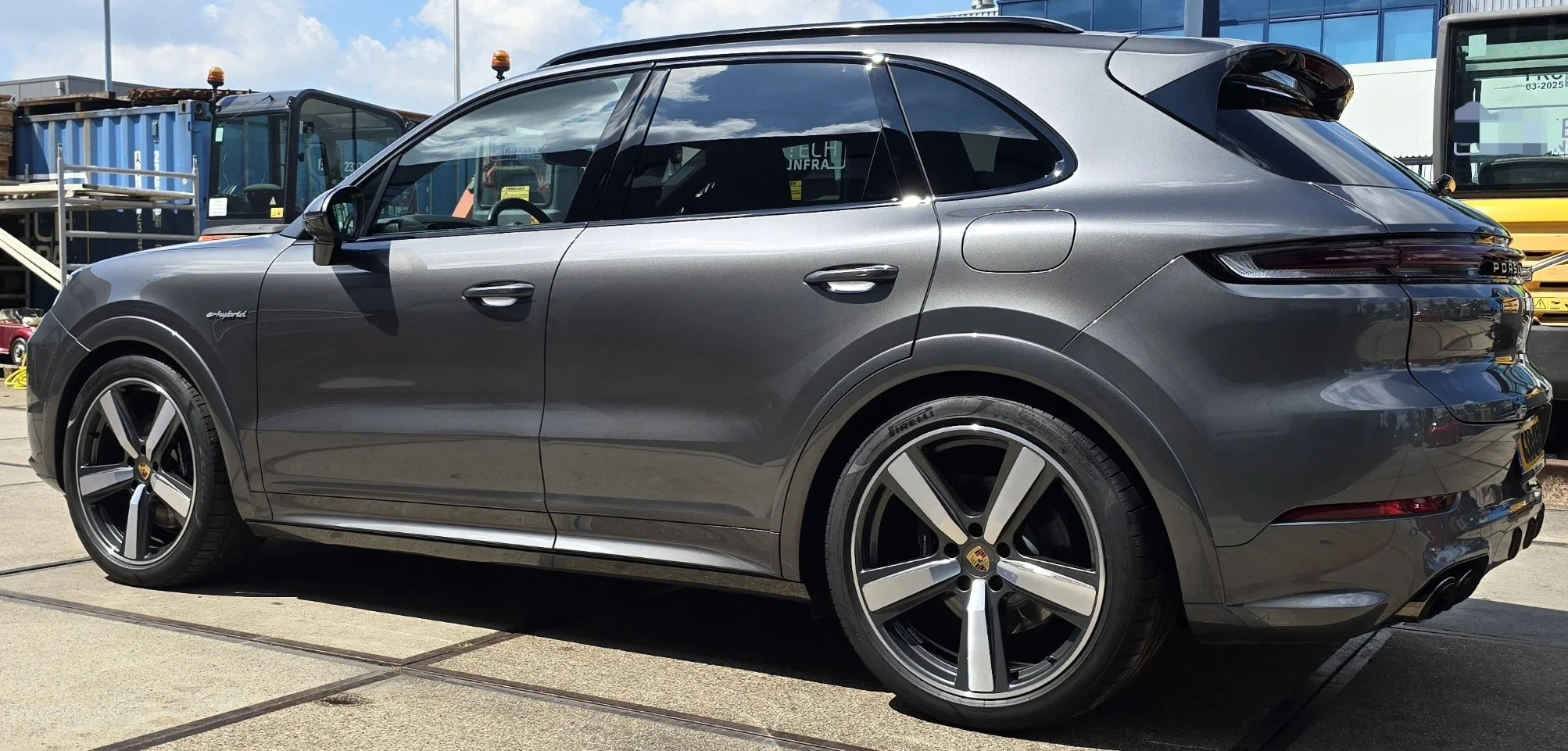 Zilverkleurige Porsche Cayenne met sportvelgen en een badge die zegt 'e-hybrid', geparkeerd op een parkeerterrein met andere voertuigen op de achtergrond.