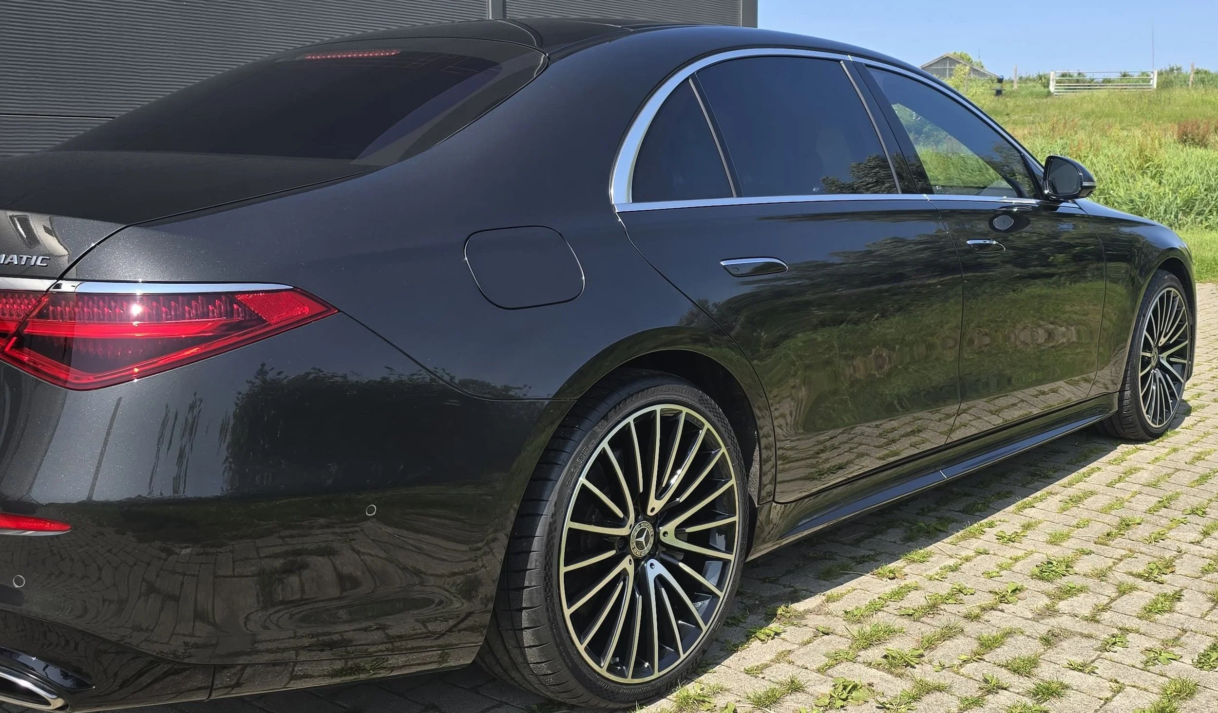 Zwarte luxe sedan auto geparkeerd op een stenen weg met groene velden en een blauwe lucht op de achtergrond.