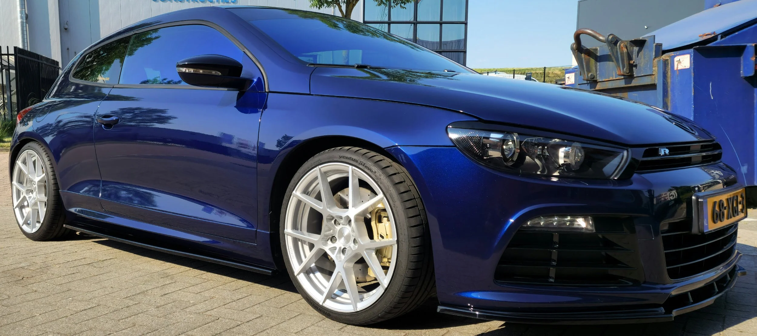 Blauwe sportauto met zilverkleurige velgen, geparkeerd op een stenen ondergrond; metalen container op de achtergrond.