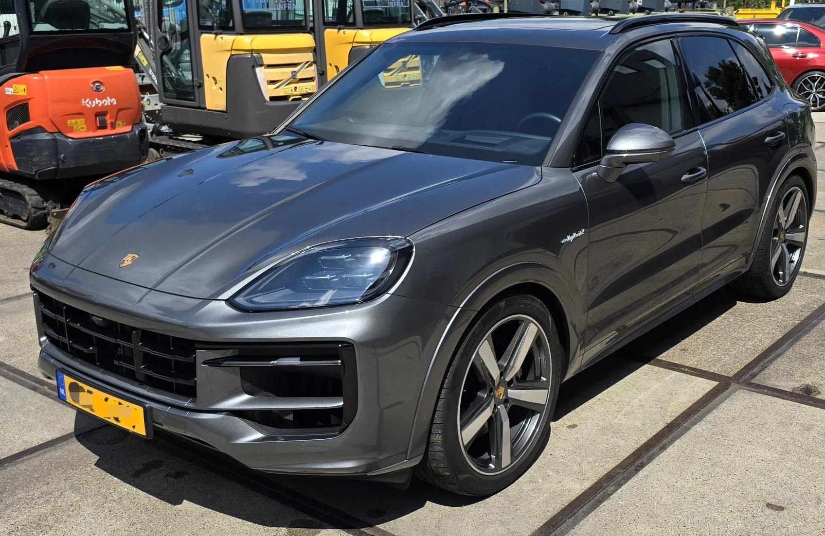 Een grijze Porsche Macan met donkere wielen, geparkeerd op een parkeerplaats naast bouwmachines en andere voertuigen.