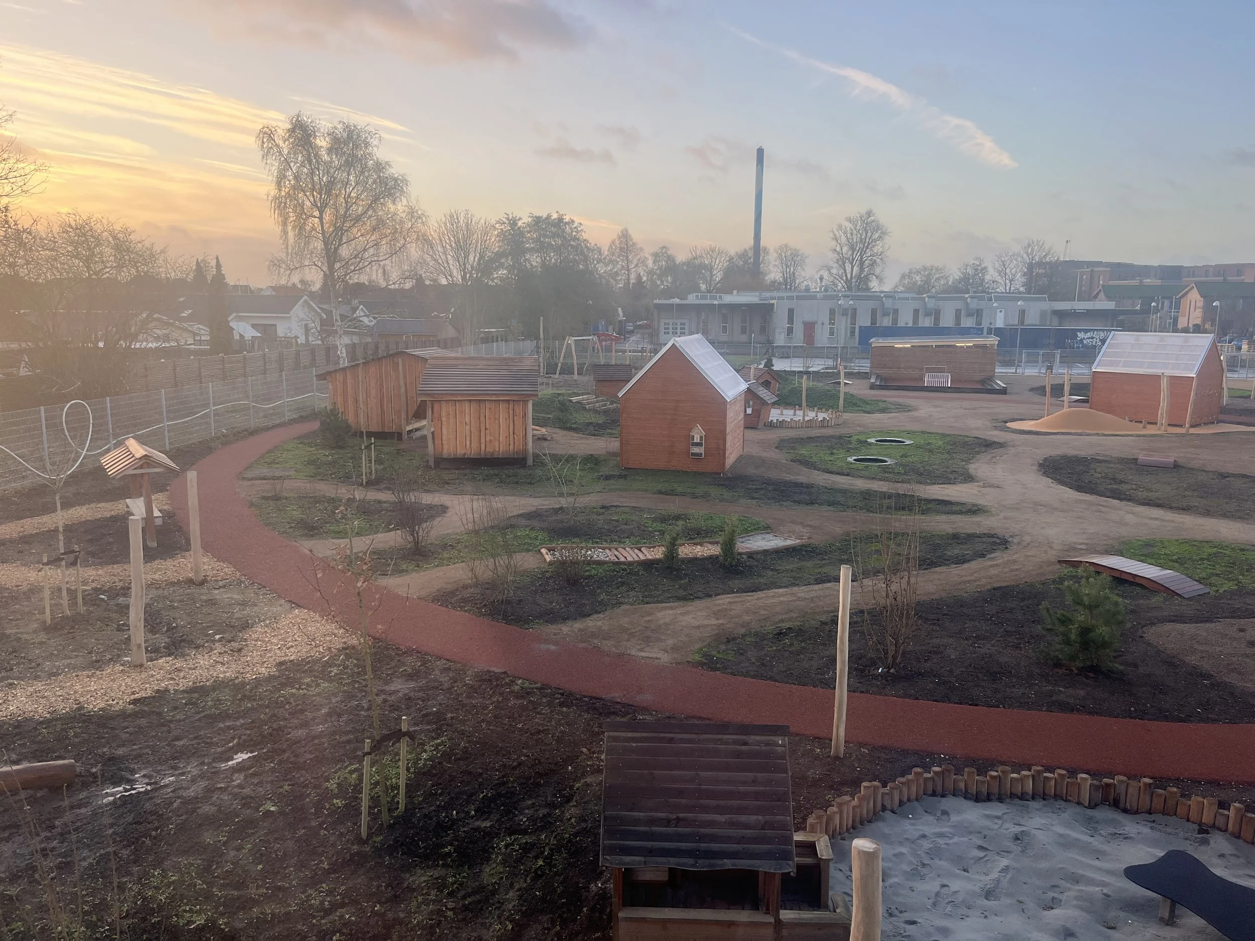 En legeplads med små træhytter, stier, legeudstyr som sandkasse og hoppepude, omgivet af træer og grønne områder under en solnedgang.