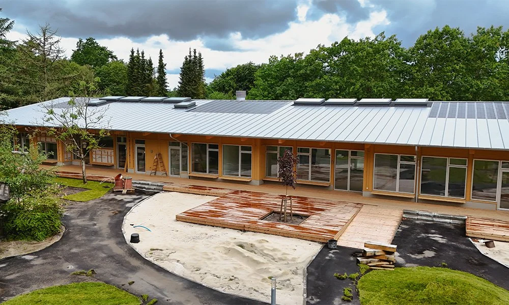 Et byggeri under opførelse med træfacader, store vinduer og et nyt tag. Der er en oprettet plante i midten af en træplatform foran huset, og der er stadig arbejde i gang med udendørsarealer.