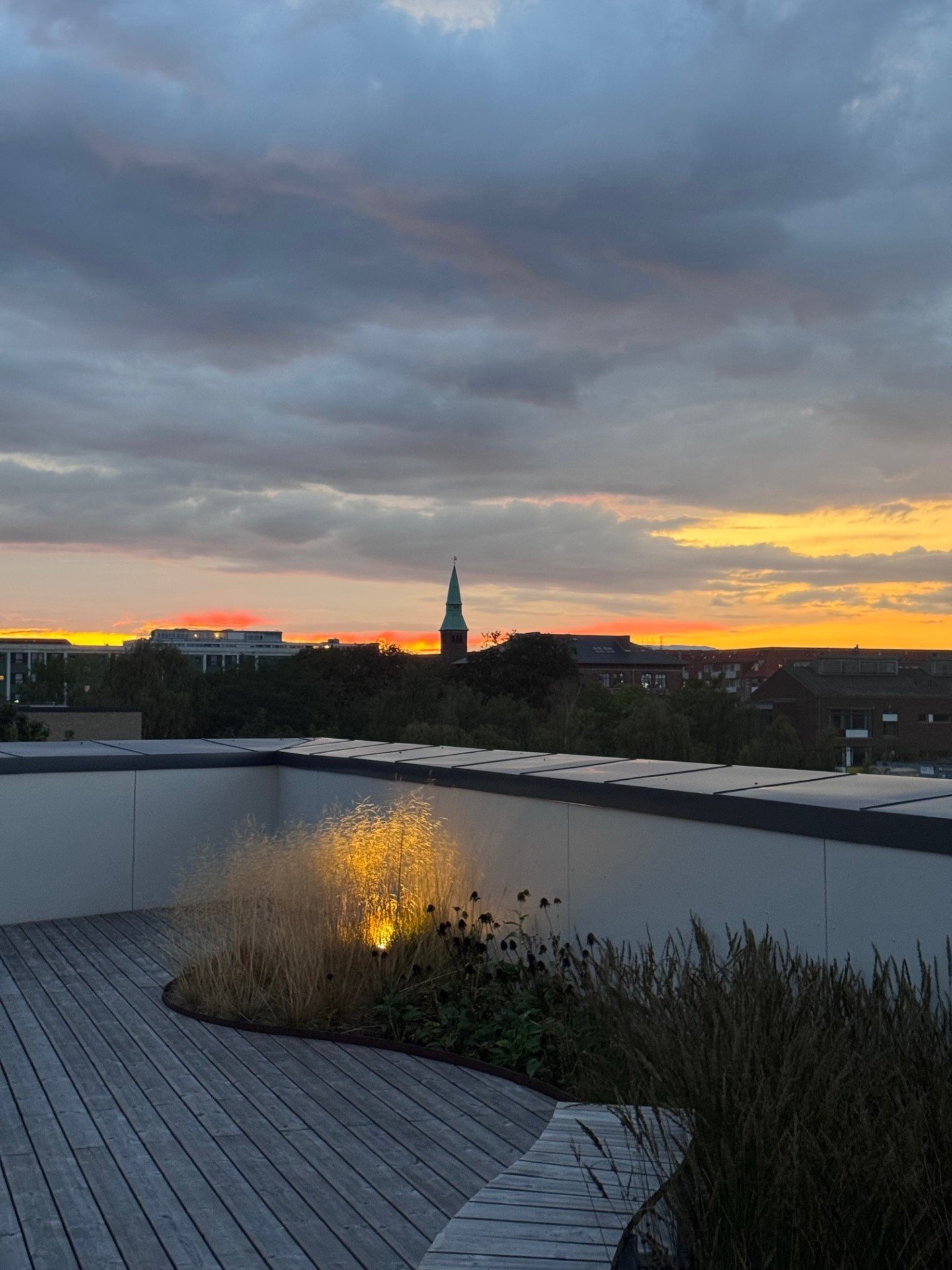 Udsigt over en by ved solnedgang med skyer på himlen, et kirkespir og en terrasse med planter