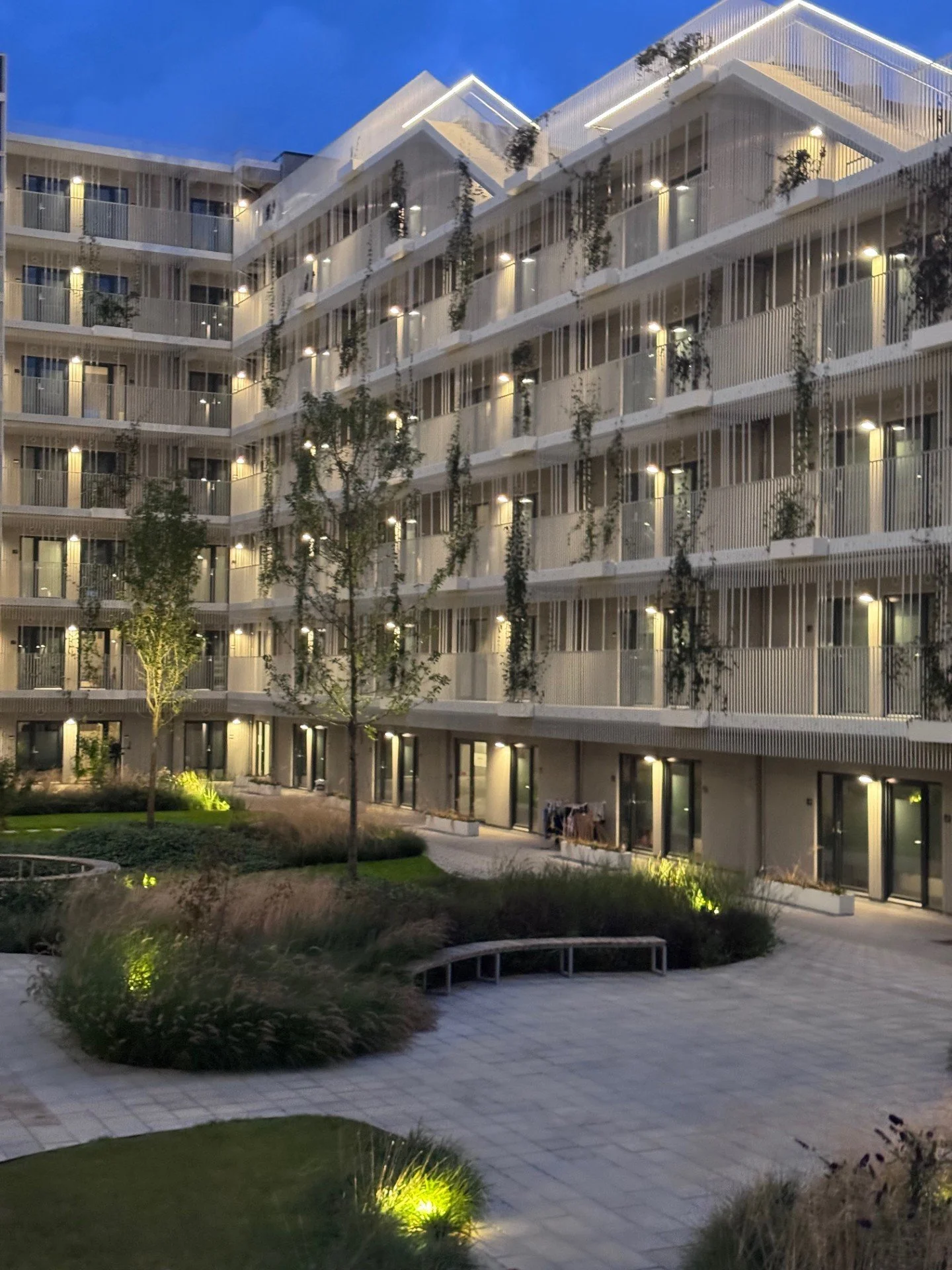 Modernt boligkompleks med udendørs verandaer og grønne områder om aftenen, oplyst af udendørs belysning.