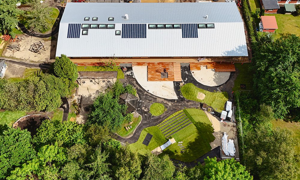 Fjerntliggende udsigt over en bygning med fladt tag, omgivet af grønt område med buskads og legeplads.