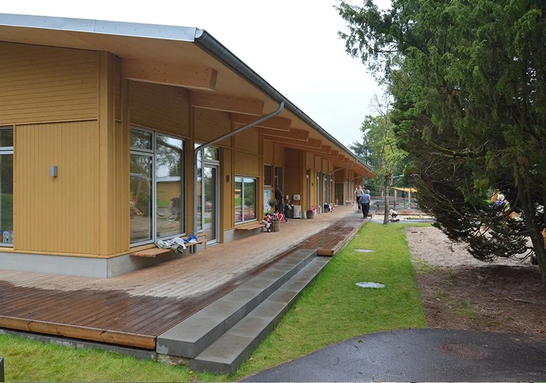 Udendørs område foran en bygning med træfacade og store vinduer, personer går rundt, grønne træer til højre.