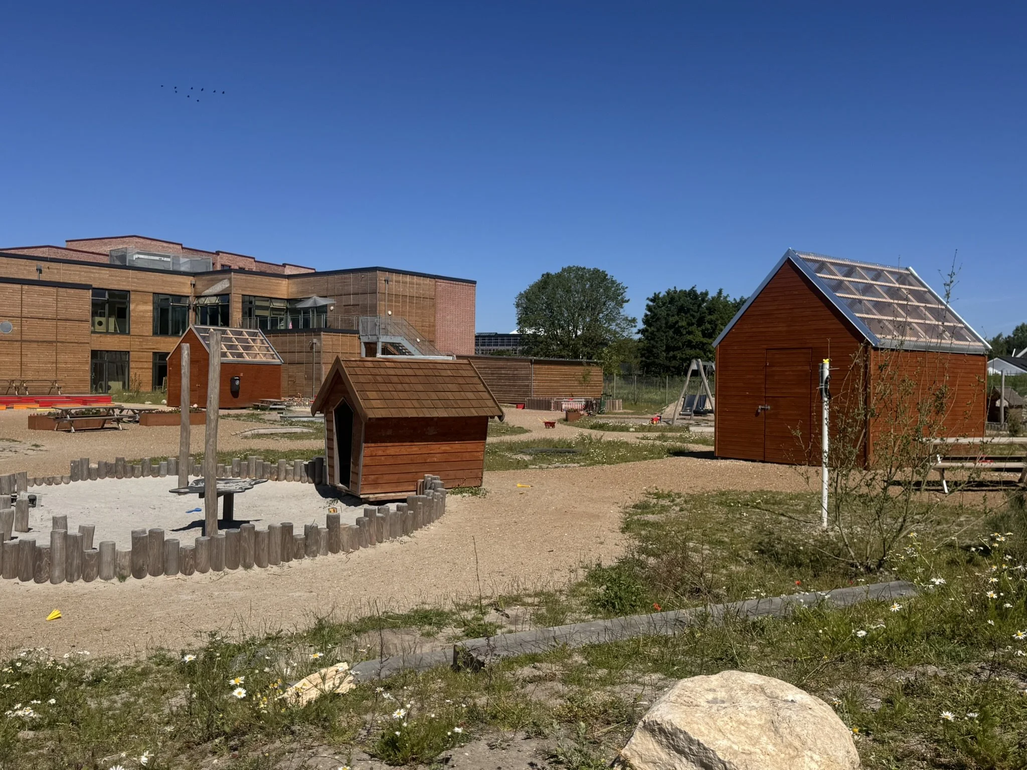 Børnelegeplads med små træhytter, sandkasse, gyngestativ og træer under en klar blå himmel.