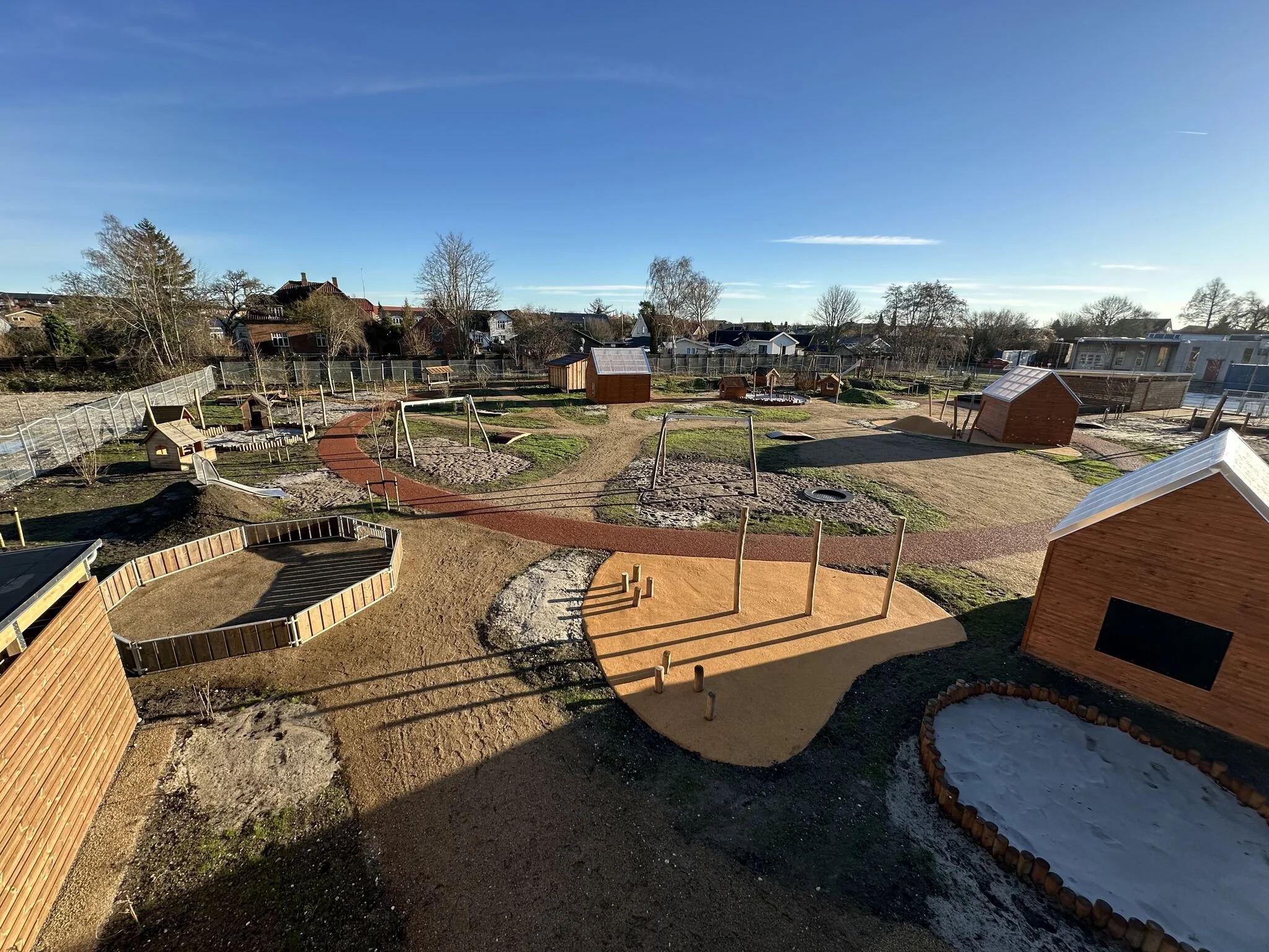 Børnelegeplads med gynger, sandkasse, små hus og plæner under en klar blå himmel.