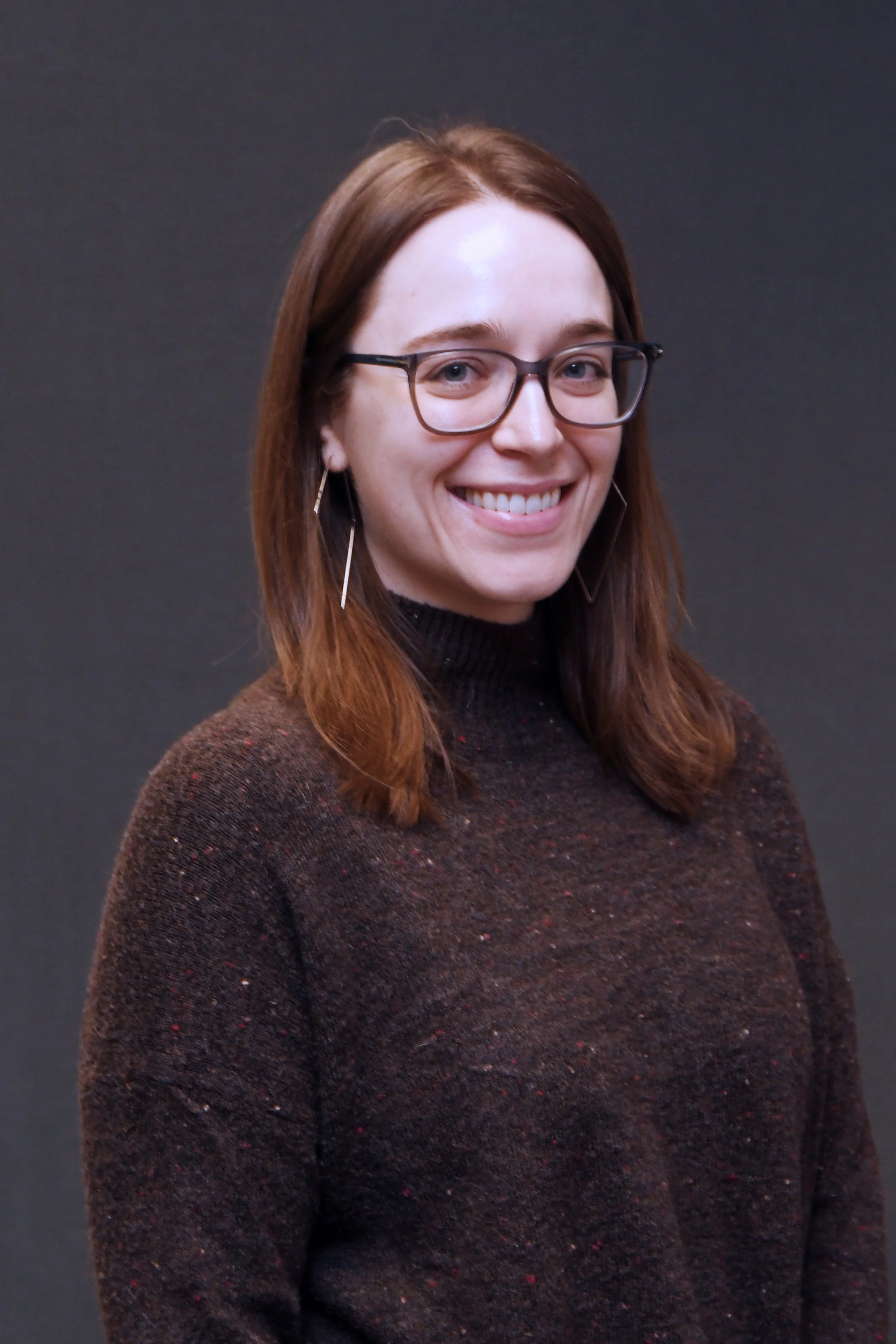 A woman with red hair and glasses smiling, wearing a dark sweater with a black background.