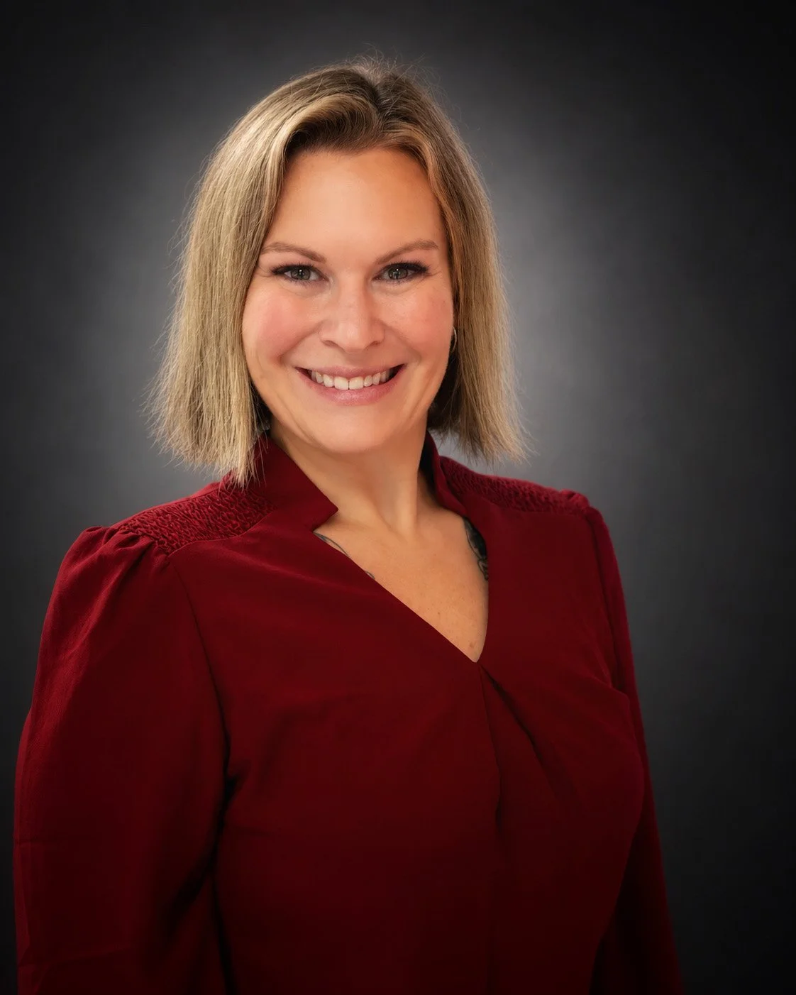 Portrait of a woman with shoulder-length blonde hair, wearing a red dress, smiling, against a dark gray gradient background.