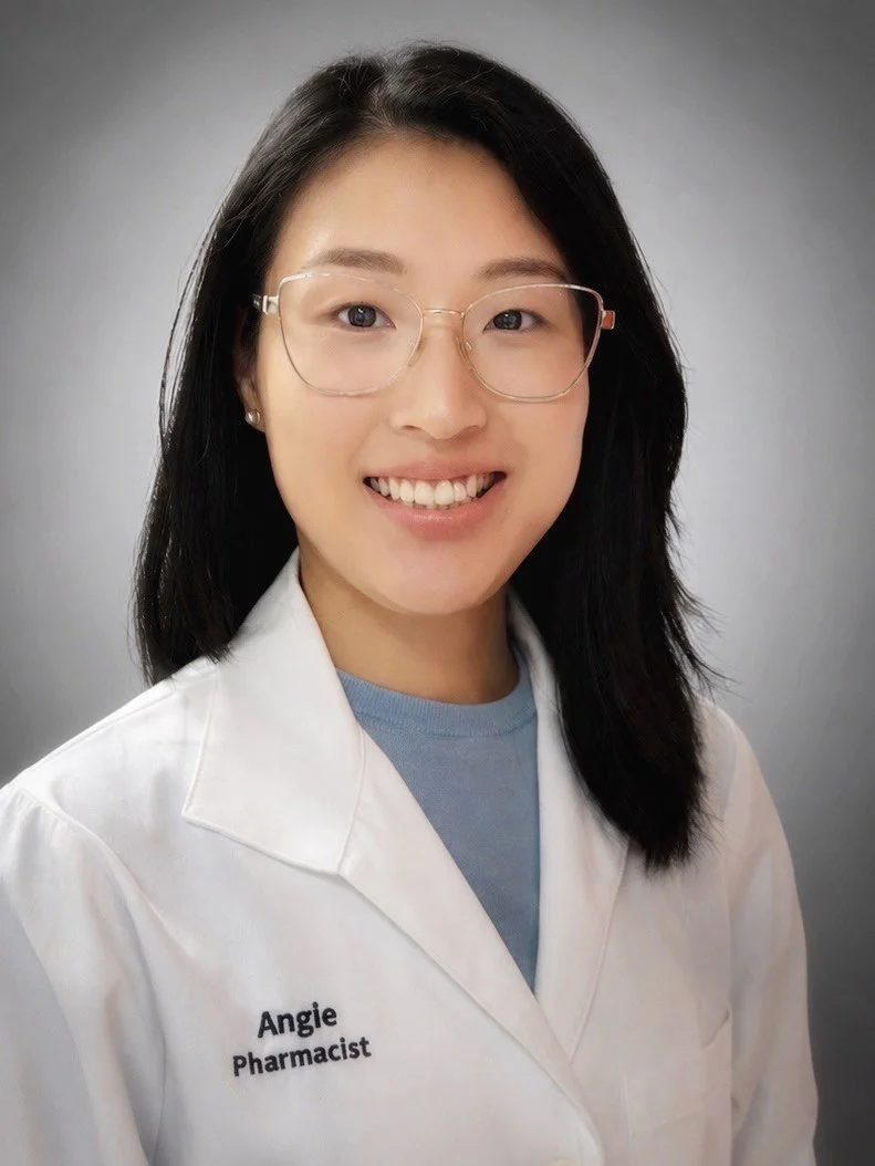 A smiling woman with long black hair, glasses, and fair skin, wearing a black blouse with a white pattern, a green cardigan, and a necklace, standing against a plain light gray background.