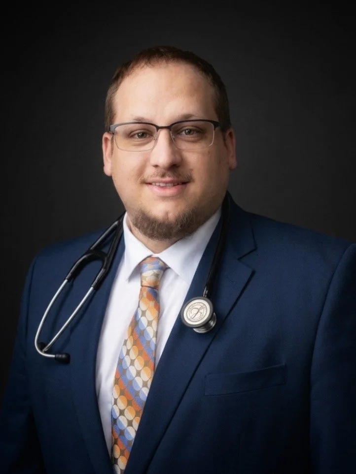 Portrait of a young male doctor wearing glasses, a navy suit, a white shirt, and a colorful patterned tie, with a stethoscope around his neck, smiling against a dark background.