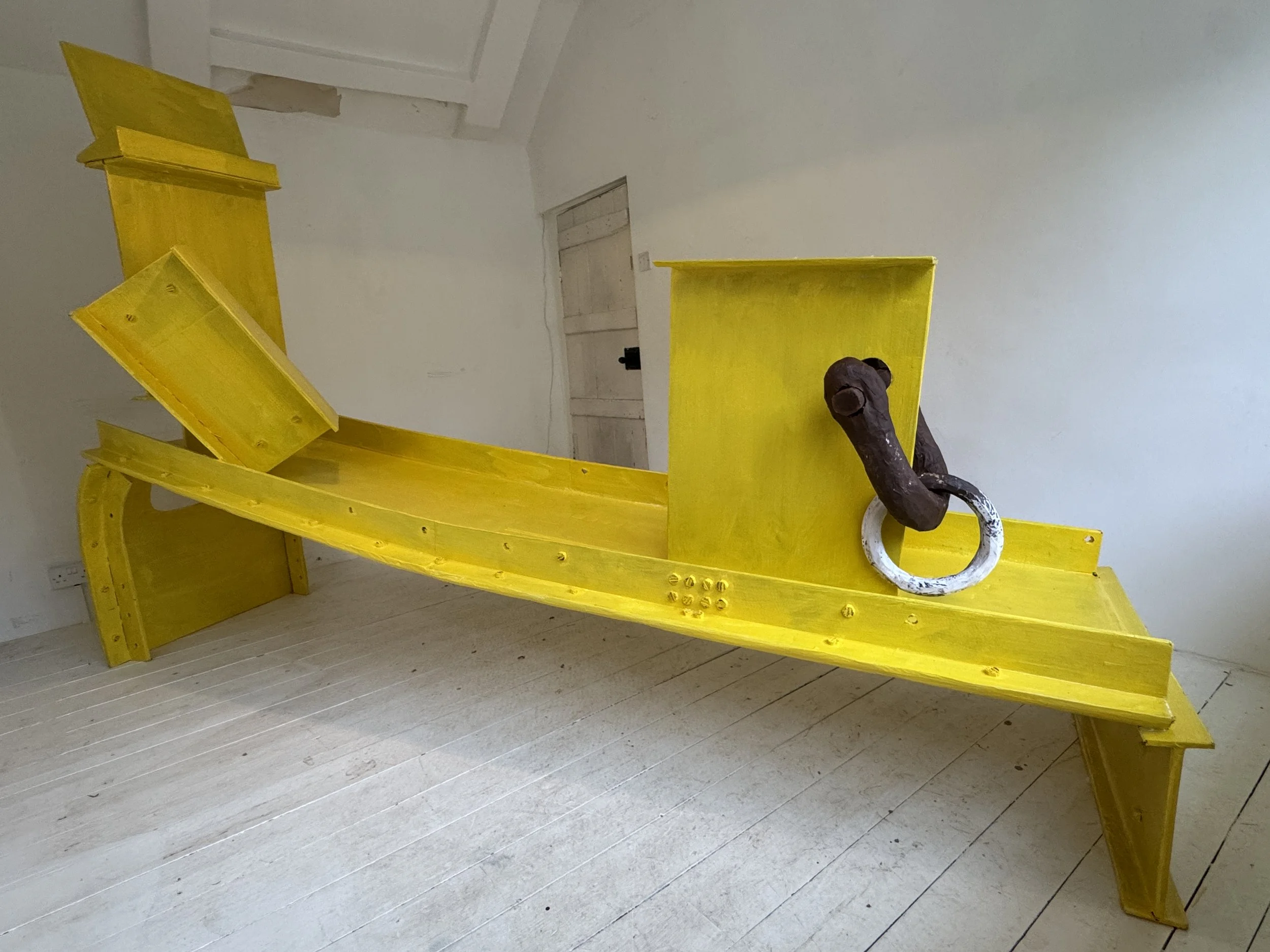 Bright yellow painted cardboard sculpture in a domestic white cottage bedroom. The form resembles Anthony Caro's work with the addition of a brown and white painted cardboard shackle and ring