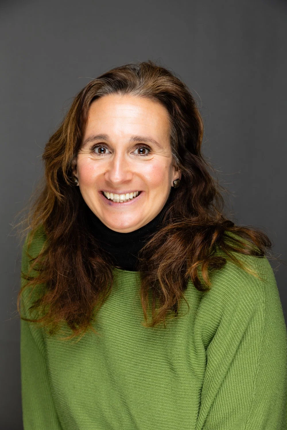 A smiling woman with long, curly brown hair, wearing a green sweater and a black turtleneck, posed against a gray background.