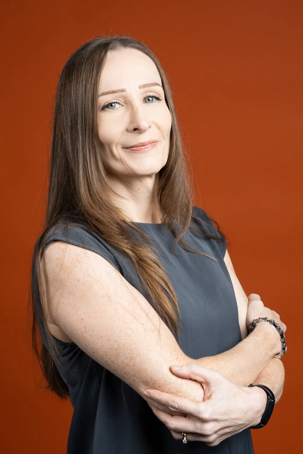 A woman with long brown hair, fair skin with freckles, wearing a dark sleeveless top, standing with arms crossed, smiling slightly, in front of an orange background.