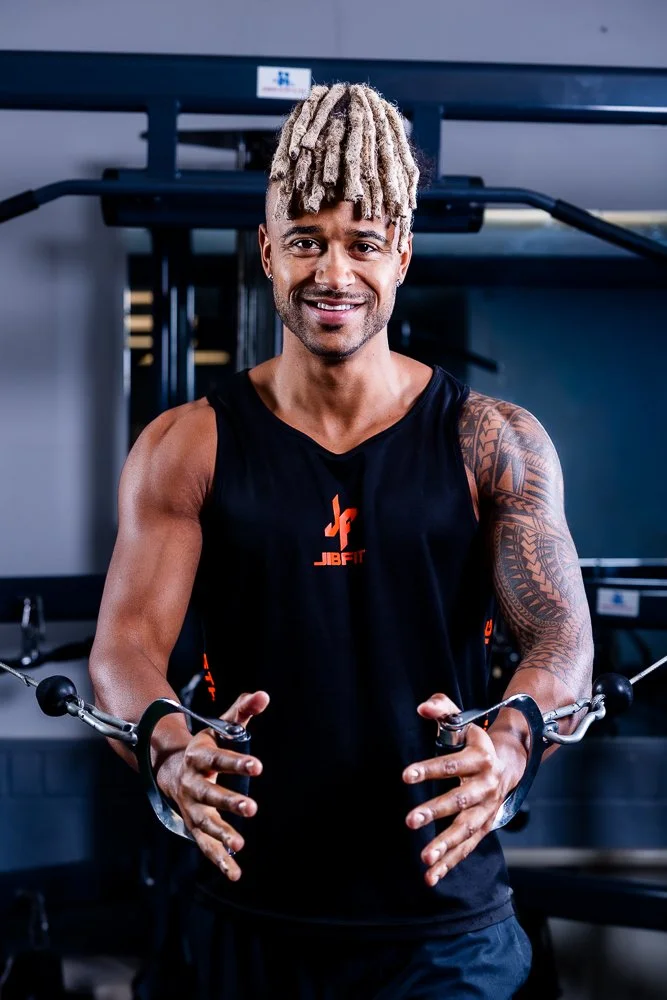 A smiling man with blond dreadlocks wearing a black sleeveless sports shirt, holding wrist weights, standing in a gym with workout equipment in the background.