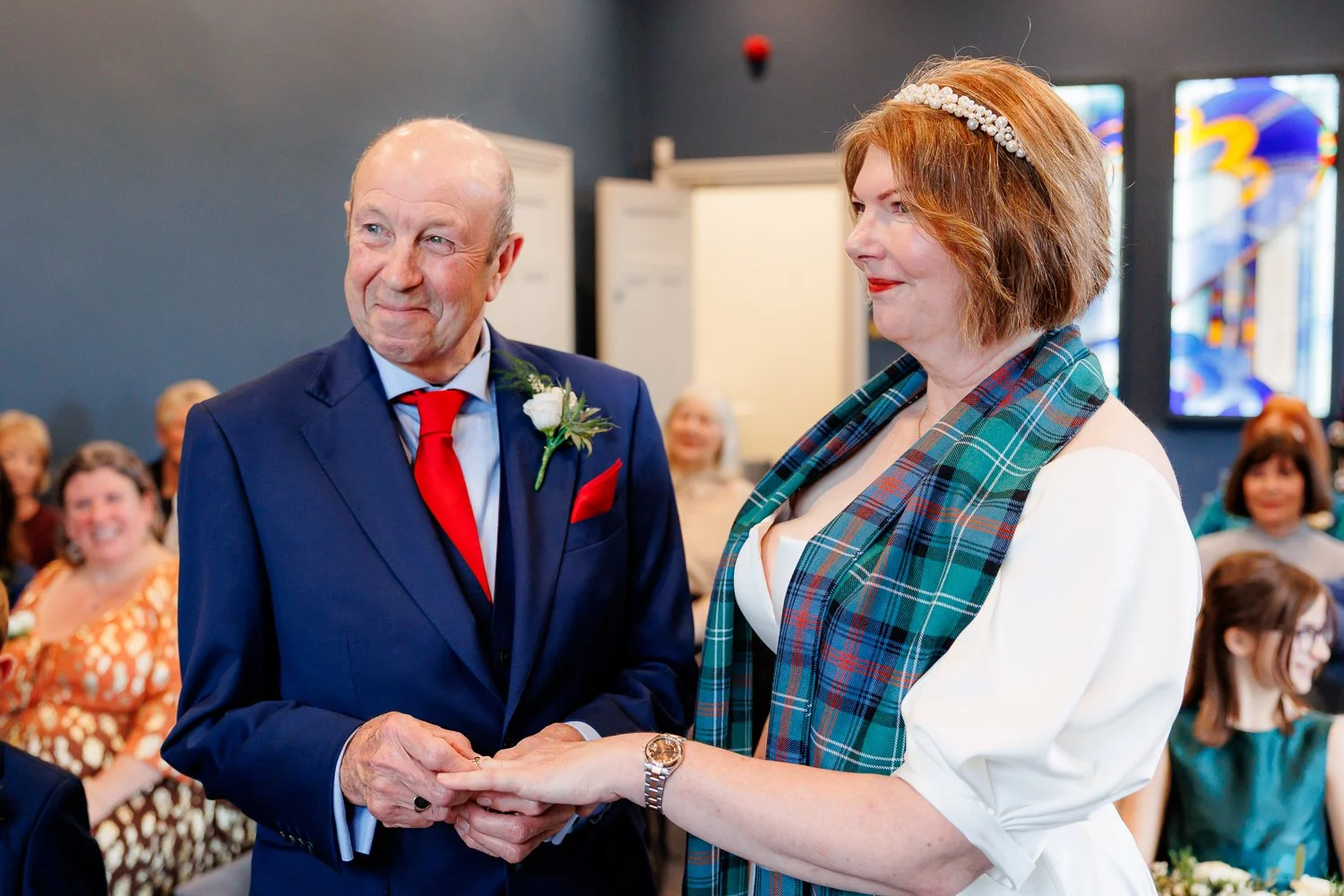A groom and bride exchanging rings during a wedding ceremony with guests watching and smiling in the background.