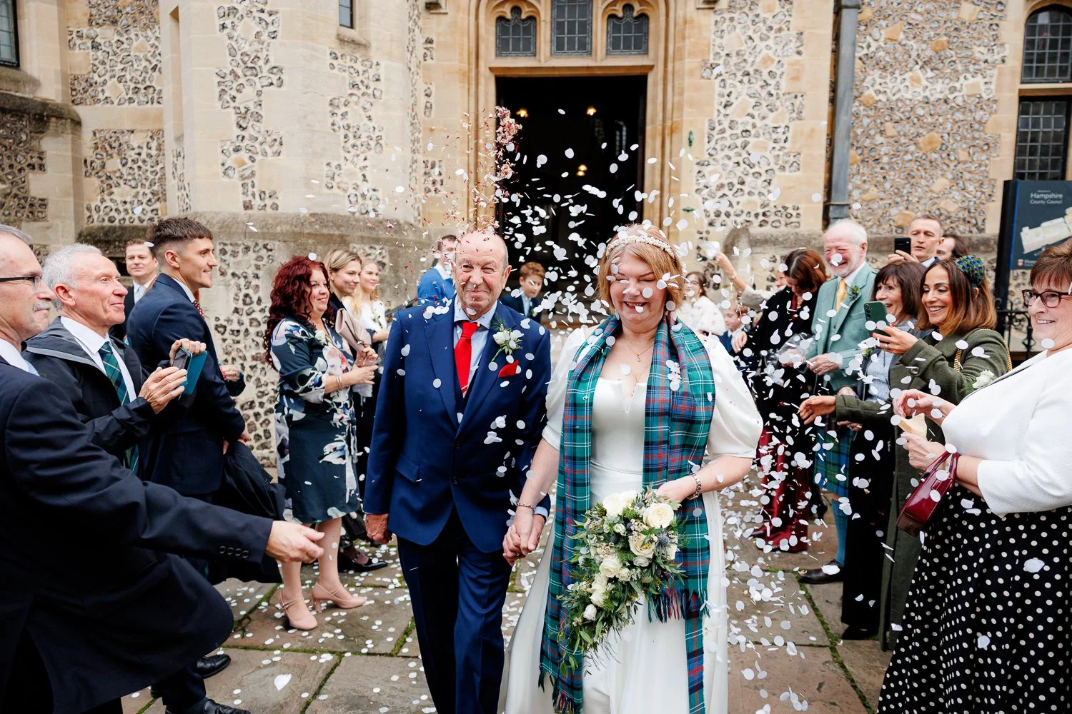 A bride and her father exiting a church with friends and family throwing confetti. The bride is holding a bouquet and wearing a white dress with a plaid scarf. The scene is joyful and celebratory.