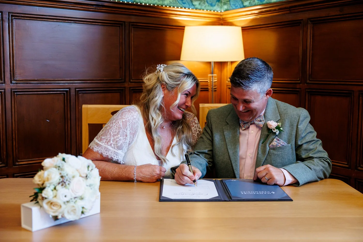 A woman in a white lace dress and a man in a light gray suit signing a marriage certificate at a wooden table. The woman has long blonde hair with a floral hair accessory, and the man has short gray hair, a floral bow tie, and a boutonniere. There is