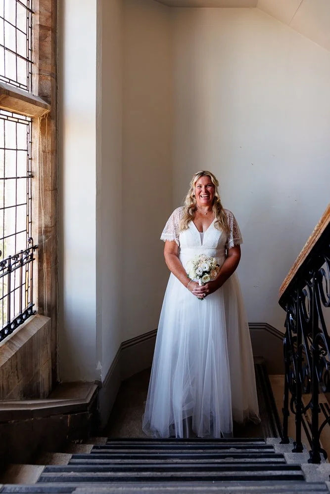 A smiling woman in a white wedding dress holding a bouquet standing on a staircase near a large window with iron bars.