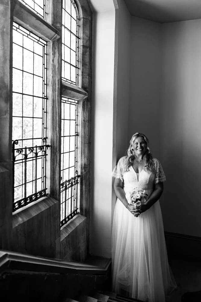 A smiling woman in a white wedding dress holding a bouquet of flowers, standing next to large decorative stained glass windows in a well-lit room.