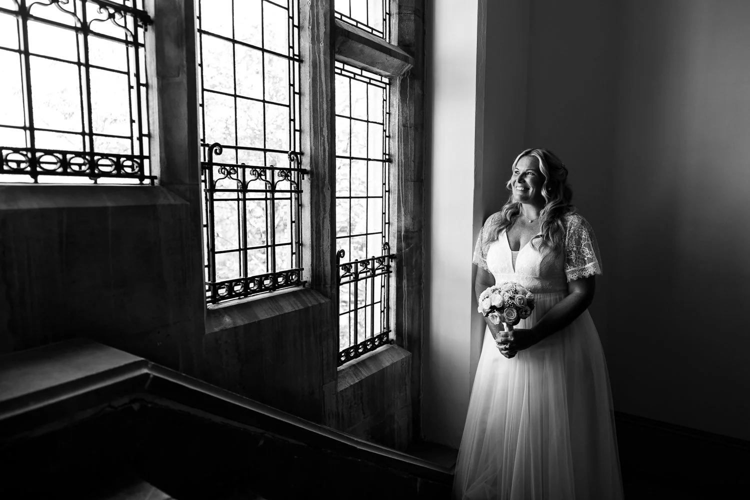 A woman in a wedding dress holding a bouquet of flowers, standing by a window with ornate iron bars, smiling as she looks outside in a black and white photograph.