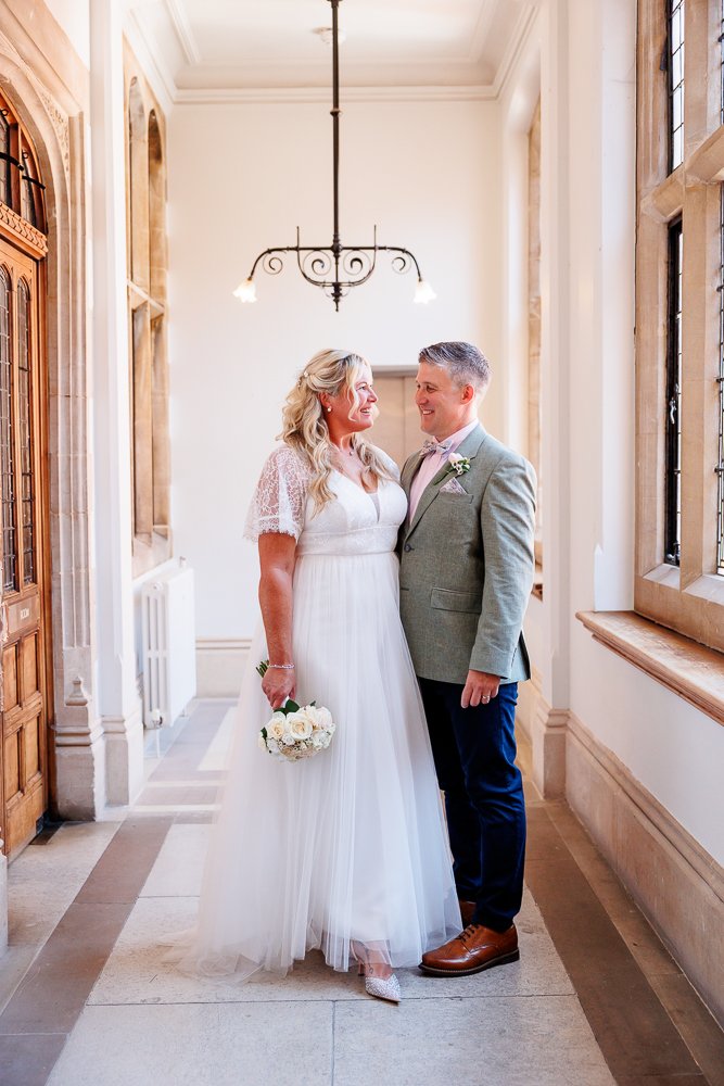 Bride and groom standing close together in a hallway with large windows and ornate light fixture, looking at each other and smiling. Bride in a white dress holding a bouquet, groom in a gray suit with a boutonnière.