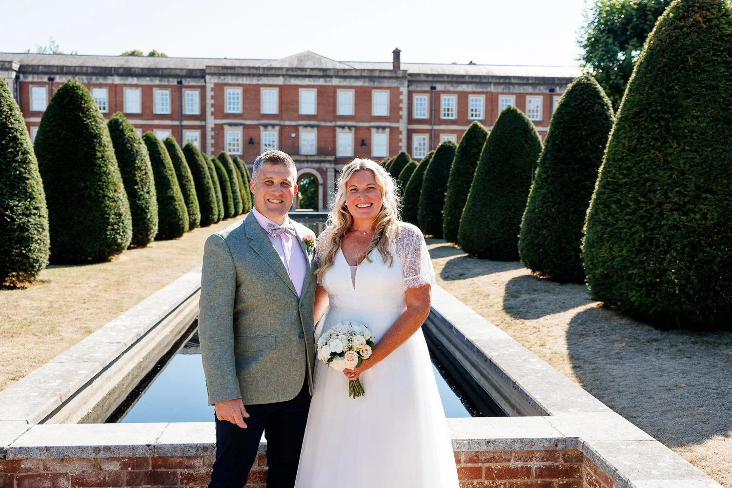 A bride and groom standing outdoors in front of a historic building with manicured trees and a fountain, smiling for a wedding photo.