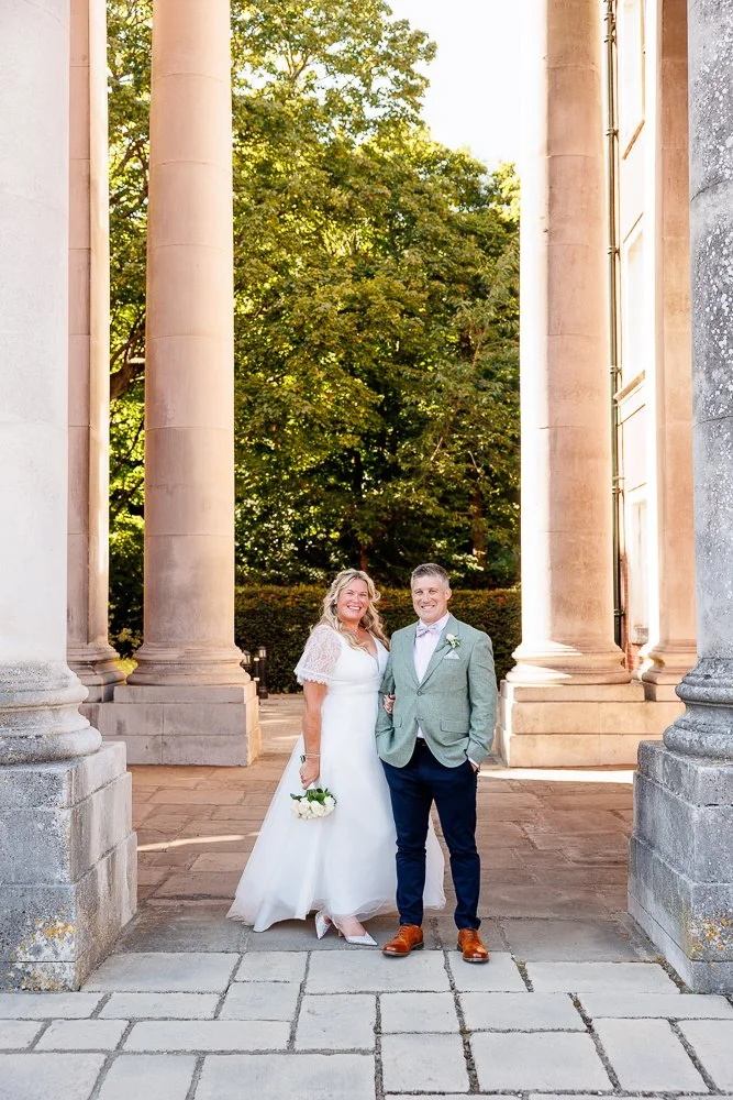 A bride and groom in wedding attire standing together outdoors, framed by large stone columns with green trees in the background.