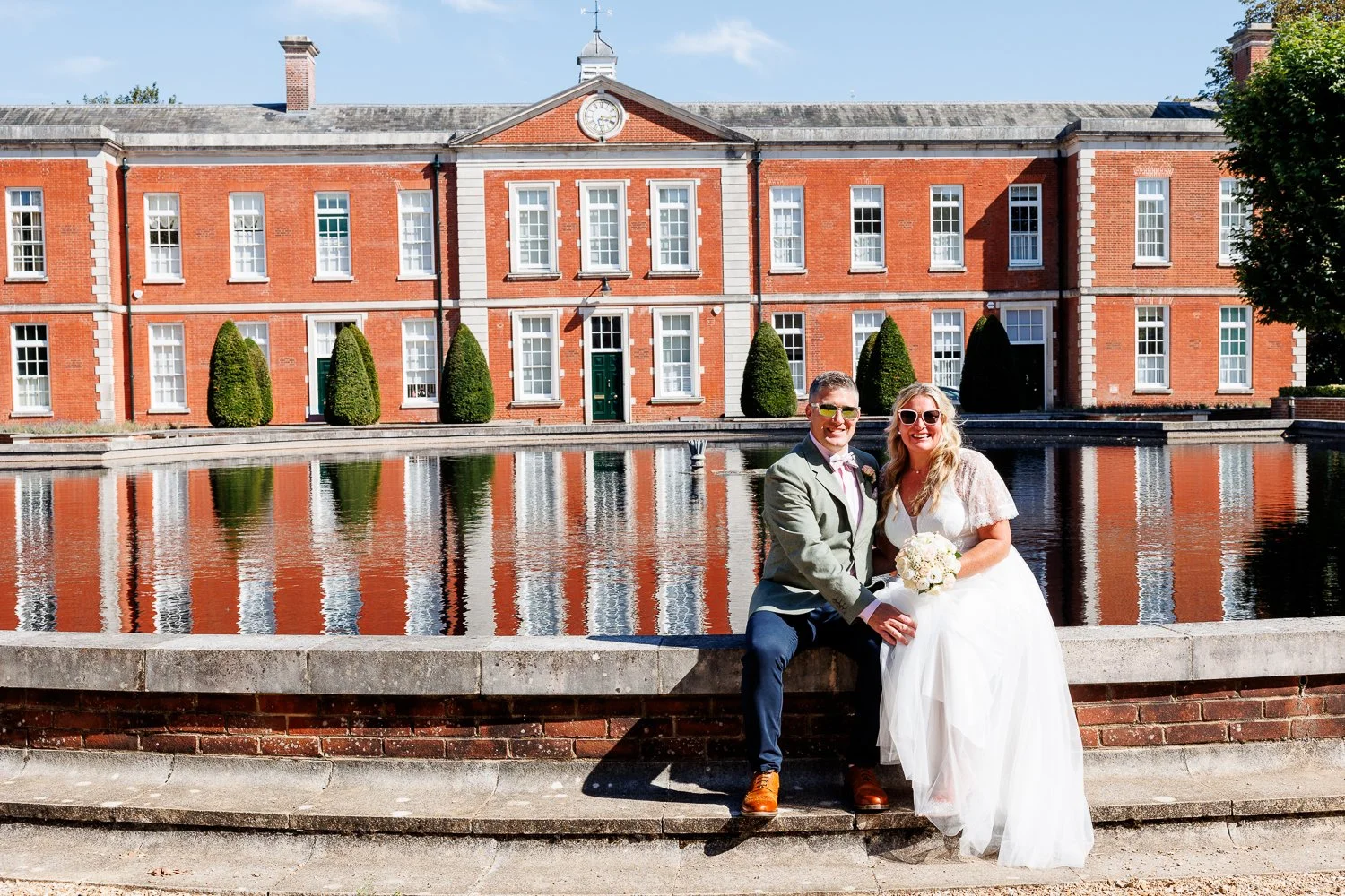 A newlywed couple sitting near a water fountain in front of a historic red brick building with white window trim and a clock, on a sunny day.