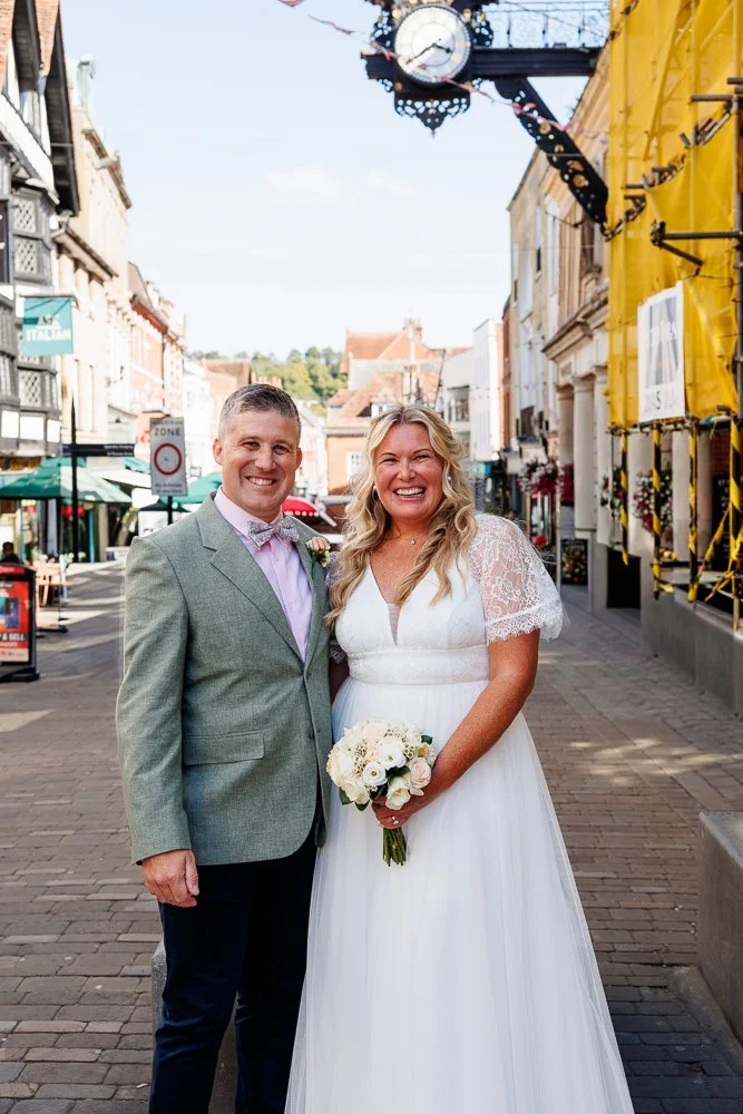 A bride and groom standing on a city street during daytime, smiling for the camera. The bride is holding a bouquet of white roses, wearing a white wedding dress with lace sleeves. The groom is dressed in a light gray suit jacket, a white shirt, and a