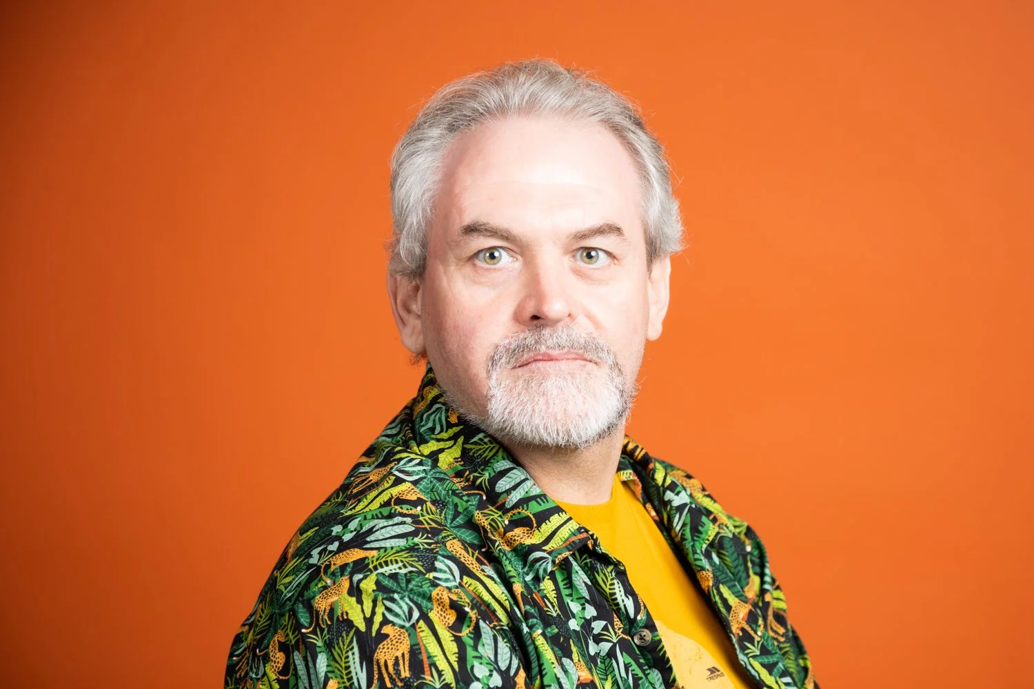 Portrait of a middle-aged man with gray hair and a beard, wearing a colorful Hawaiian shirt and yellow T-shirt, against an orange background.