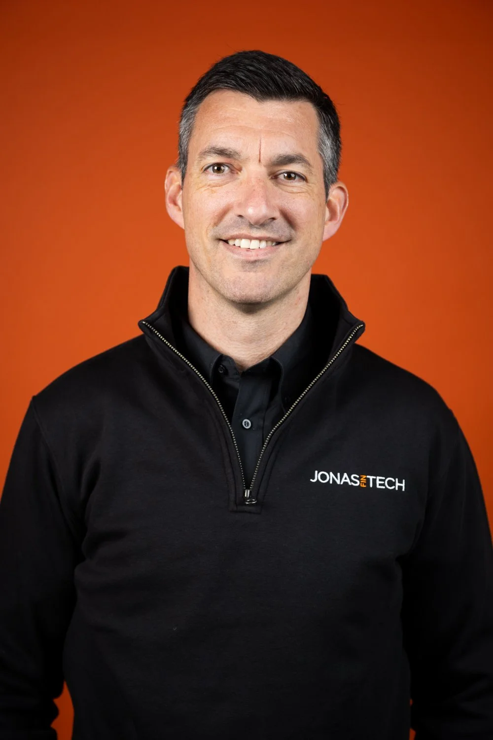 A man with short dark hair smiling, wearing a black jacket with a zip and 'JONAS IN TECH' embroidered on the chest, standing against an orange background.