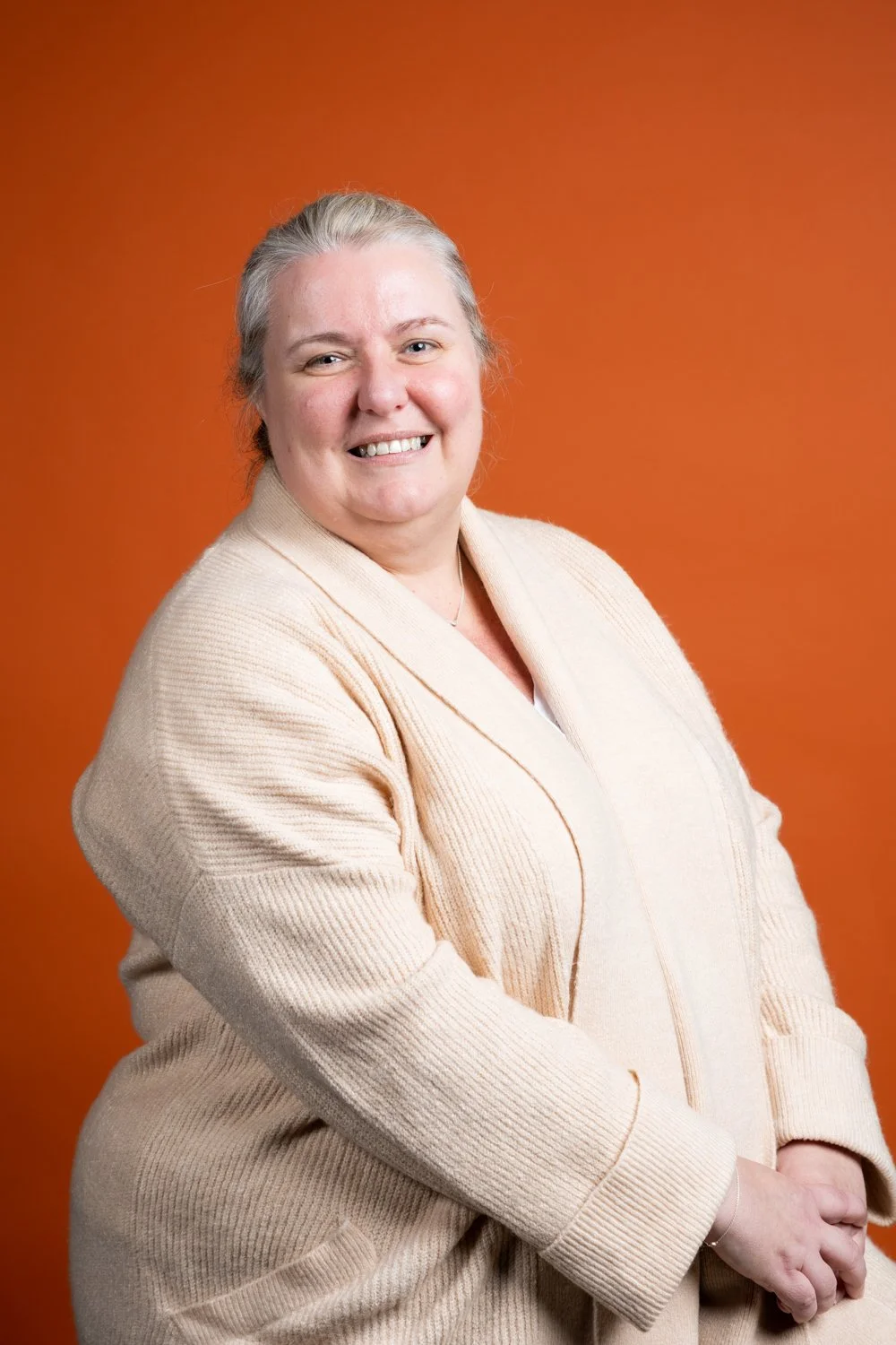 A smiling woman with gray hair in a beige cardigan against an orange background.