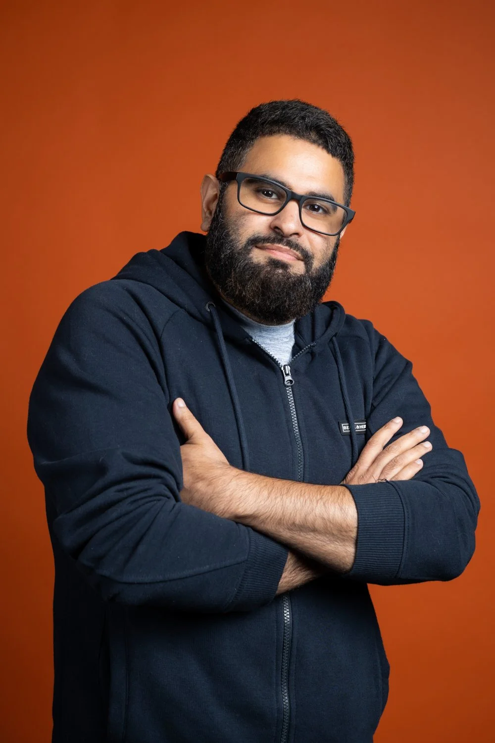 A man with glasses and a beard wearing a black zip-up hoodie, crossing his arms, against an orange background.