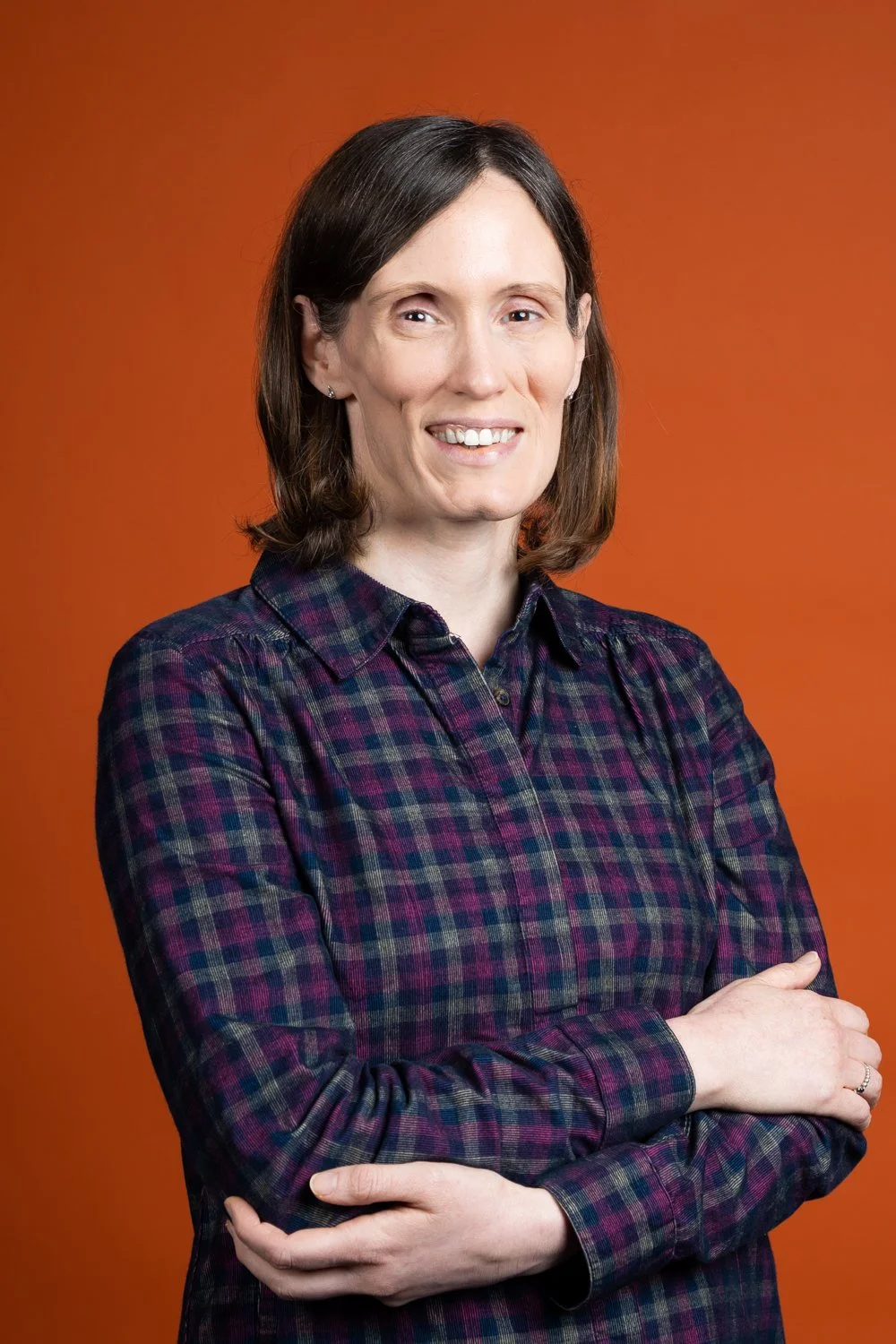 A woman with shoulder-length brown hair, wearing a purple and blue plaid shirt, standing with crossed arms and smiling against an orange background.