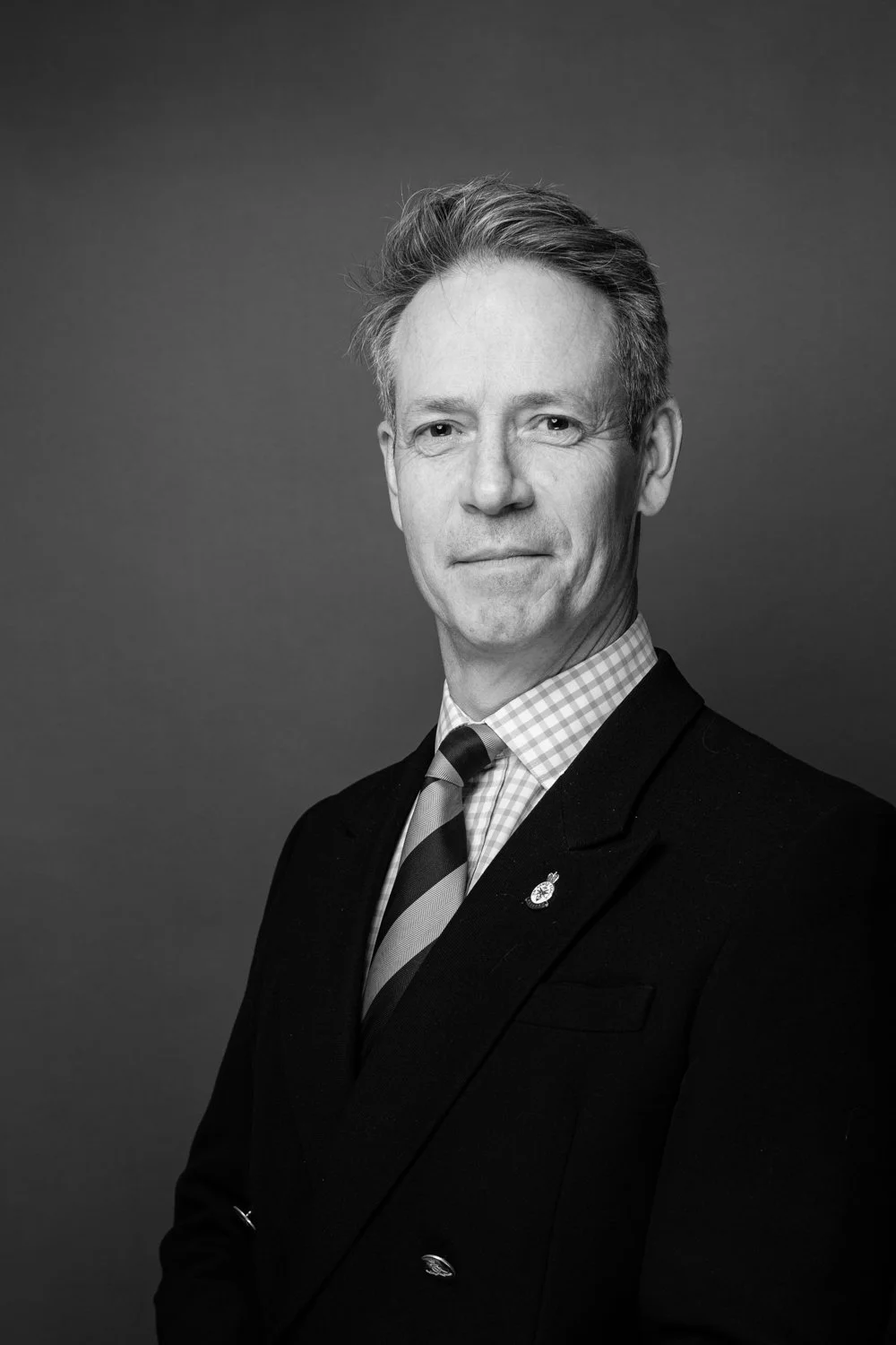 Black and white portrait of a middle-aged man with short, light-colored hair, wearing a dark suit, checkered shirt, and striped tie, against a plain background.