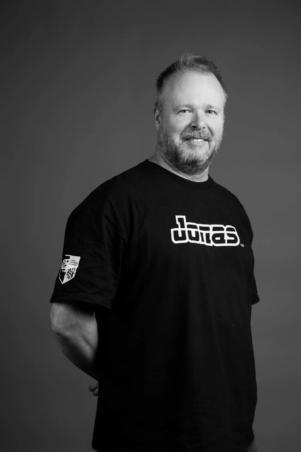 Black and white portrait of a middle-aged man smiling, wearing a black t-shirt with a graphic logo and an emblem on the sleeve, against a plain background.