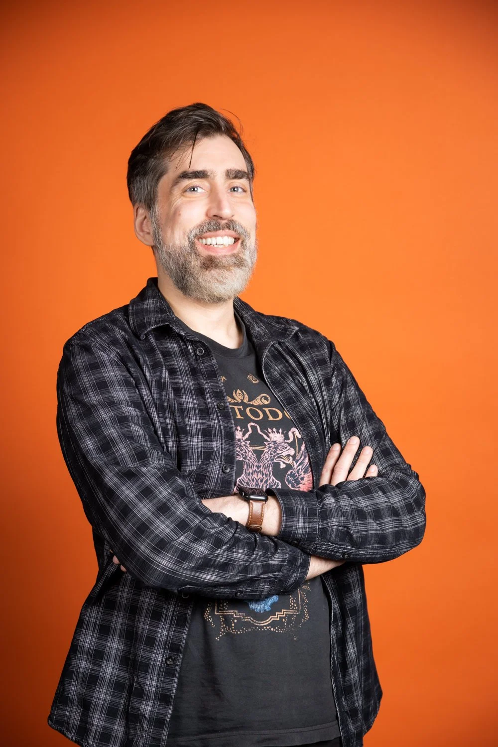Man with gray beard smiling and crossing arms, standing against an orange background.