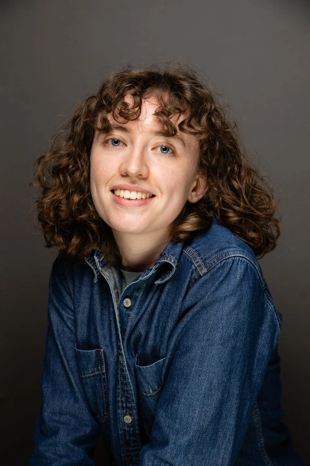 A young woman with curly brown hair, light skin, and blue eyes, smiling, wearing a denim jacket against a plain gray background.