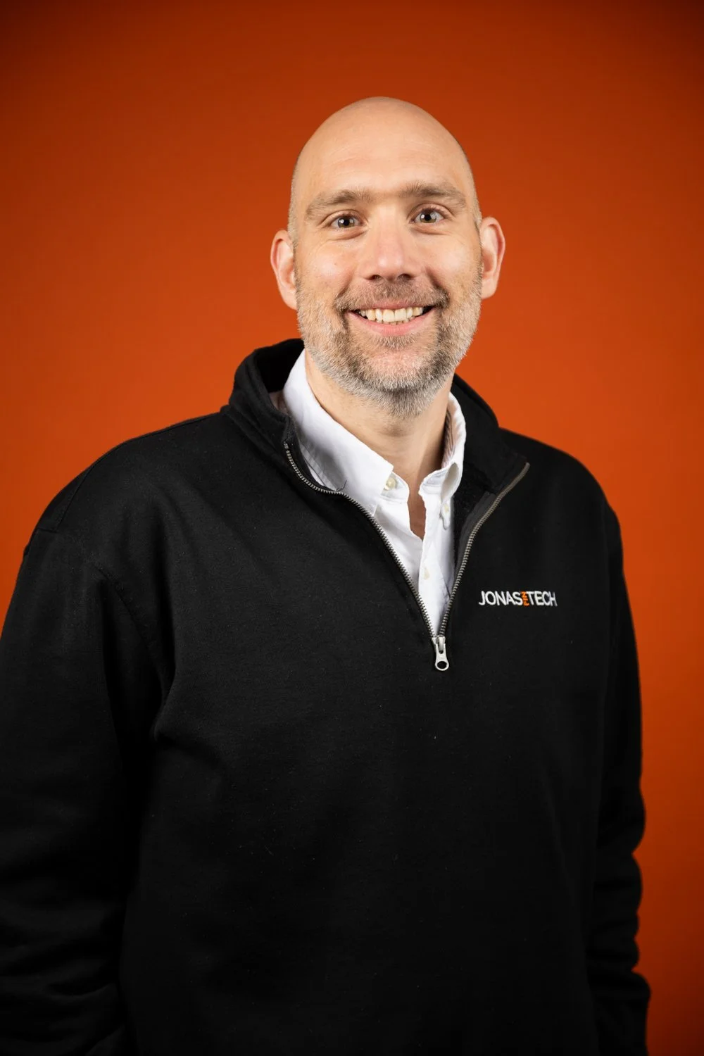 A smiling man with a beard and bald head wearing a white dress shirt and a black Jonasz Tech zip-up fleece jacket standing against an orange background.