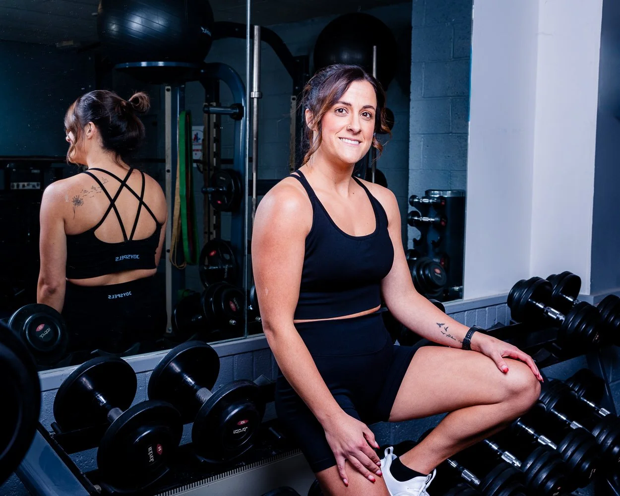 A woman sitting on a bench in a gym surrounded by dumbbells, smiling at the camera with workout equipment and mirrors in the background.