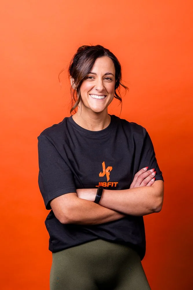 A woman smiling with arms crossed, wearing a black JB FIT T-shirt, olive green pants, and standing against an orange background.
