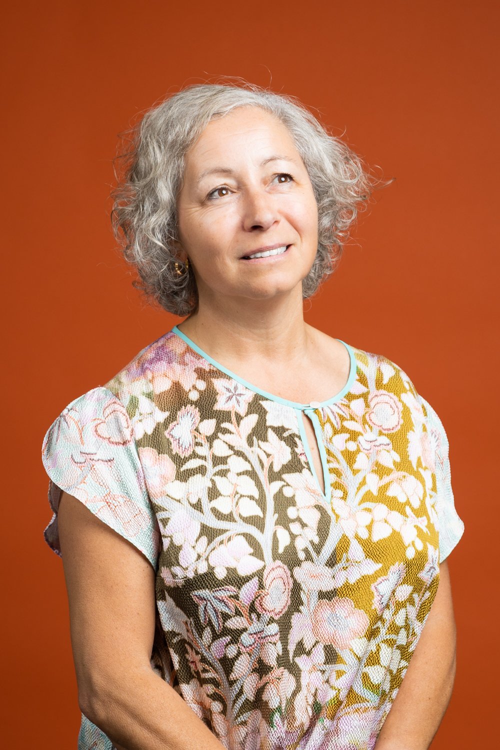 A woman with curly gray hair wearing a floral patterned top looks slightly upward against an orange background.